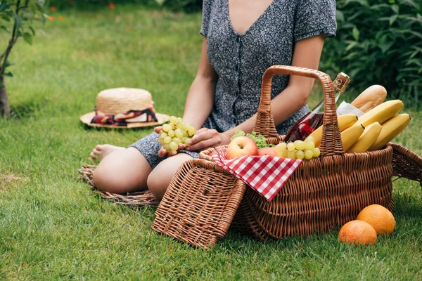 Cropped image of woman sitting on green grass at picnic and holding grapes — Stock Photo