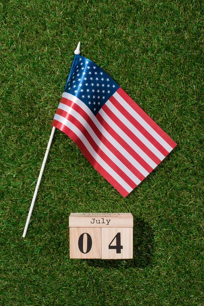 Vista superior del asta de la bandera americana y calendario de madera con fecha de 4 de julio en hierba verde, concepto de día de independencia de América - foto de stock