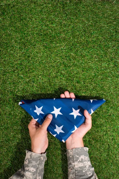 Vista parcial del soldado en uniforme militar sosteniendo la bandera doblada en las manos sobre hierba verde, concepto de fiesta del 4 de julio - foto de stock