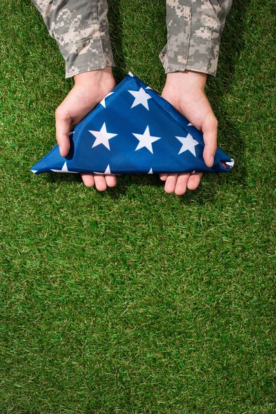 Partial view of soldier in military uniform holding folded flag in hands on green grass, 4th july holiday concept — Stock Photo