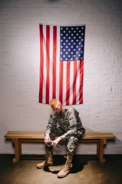 Red hair american soldier sitting on wooden bench with flag on white brick wall behind, americas independence day concept — Stock Photo