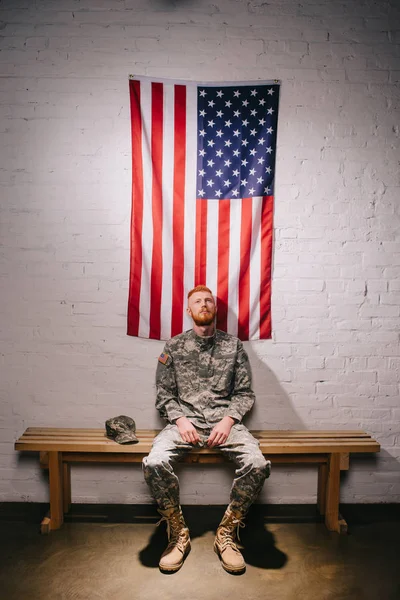 Cabello rojo soldado americano sentado en banco de madera con bandera en la pared de ladrillo blanco detrás, concepto de día de independencia de América - foto de stock