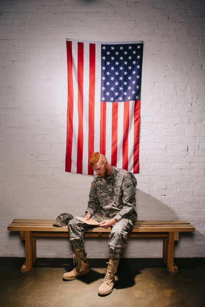 Pelo rojo soldado americano con carta sentado en banco de madera con bandera en pared de ladrillo blanco detrás, concepto de día de independencia de América - foto de stock