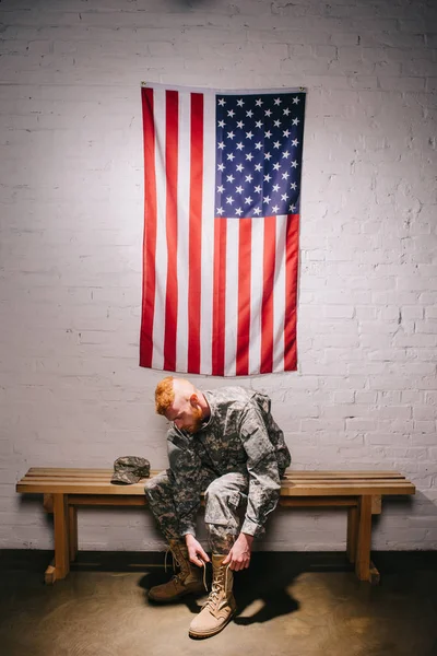 Soldado americano en uniforme militar atando cordones de zapatos con bandera en pared de ladrillo blanco detrás, concepto de fiesta del 4 de julio - foto de stock