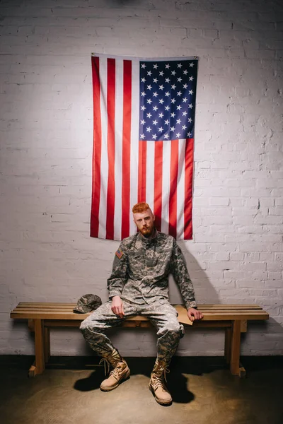 Soldat en uniforme militaire avec lettre assise sur un banc en bois avec drapeau américain sur un mur de briques blanches derrière, concept de vacances du 4 juillet — Photo de stock