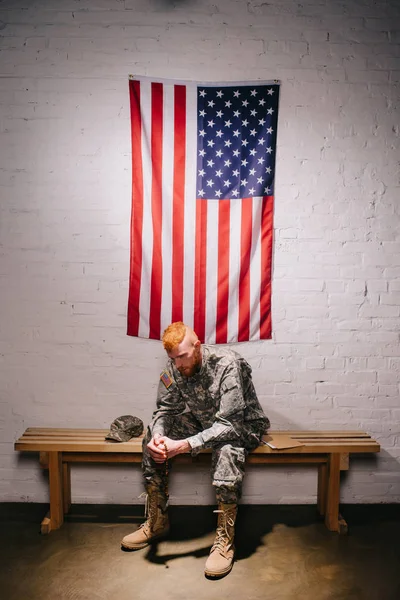Soldado en uniforme militar sentado en banco de madera con bandera americana en la pared de ladrillo blanco detrás, 4 julio concepto de vacaciones - foto de stock