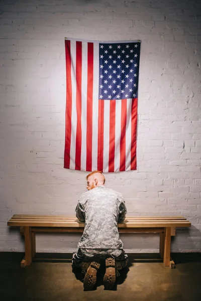 Rear view of soldier in military uniform praying with american flag on white brick wall, americas independence day concept — Stock Photo
