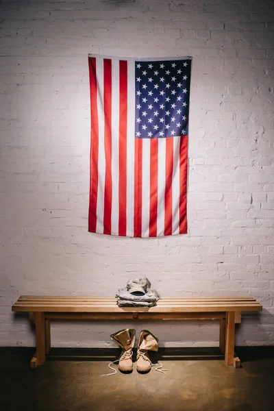 Close up view of american flag hanging on white brick wall and arranged military uniform on wooden bench — Stock Photo