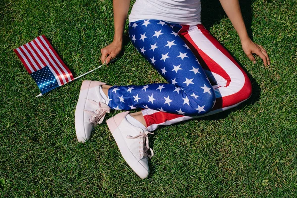 Часткове зору того, жінка з флагштока в leggins з американським прапором візерунком, відпочиваючи на зеленій галявині, americas незалежності день свято концепції — стокове фото