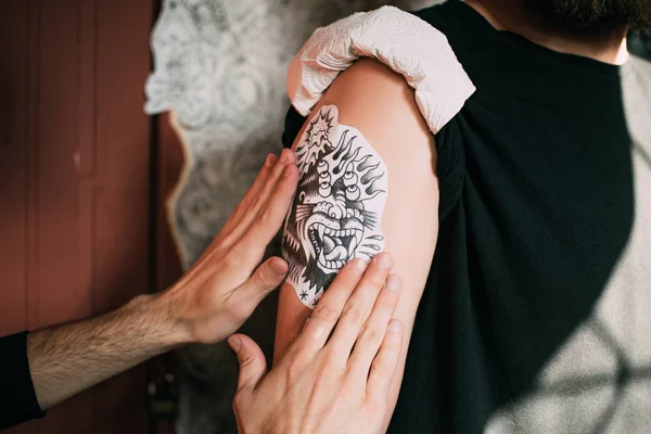 Colpo ritagliato dell'artista del tatuaggio che trasferisce lo schizzo del tatuaggio sulla spalla nel salone del tatuaggio — Foto stock