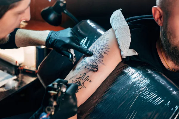 Foco seletivo do tatuador em luvas com máquina de tatuagem trabalhando na tatuagem no ombro no salão — Fotografia de Stock