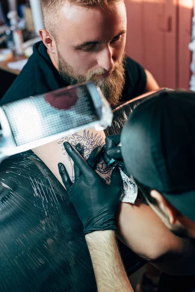 Selektiver Fokus des Tätowierers in Handschuhen mit Tätowiermaschine, die im Salon an der Tätowierung auf der Schulter arbeitet — Stockfoto