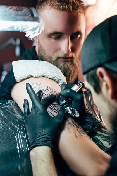 Селективный фокус татуировщика в перчатках с тату-машиной, работающей на татуировке на плече в салоне — стоковое фото