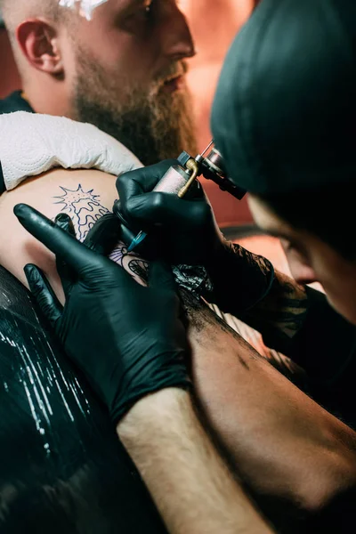Foco seletivo do tatuador em luvas com máquina de tatuagem trabalhando na tatuagem no ombro no salão — Fotografia de Stock