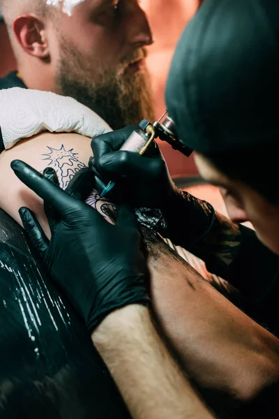 Selektiver Fokus des Tätowierers in Handschuhen mit Tätowiermaschine, die im Salon an der Tätowierung auf der Schulter arbeitet — Stockfoto