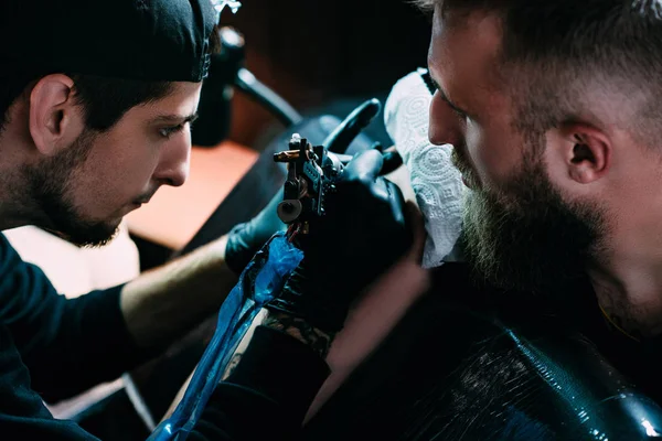 Side view of tattoo artist in gloves with tattoo machine working on tattoo on shoulder in salon — Stock Photo