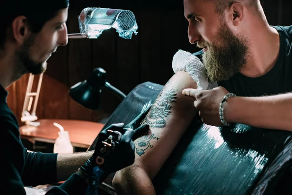 Side view of tattoo artist in gloves with tattoo machine working on tattoo on shoulder in salon — Stock Photo