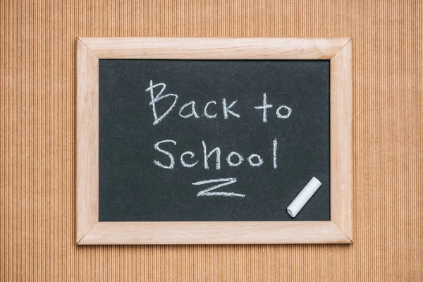 Top view of blackboard with back to school inscription and piece of chalk on brown background — Stock Photo