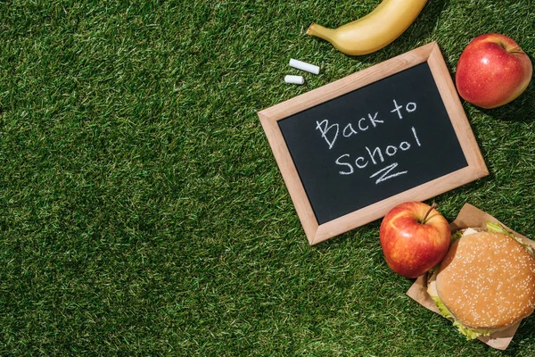 Flat lay with blackboard with back to school lettering, pieces of chalk, banana, apples and burger on green grass — Stock Photo