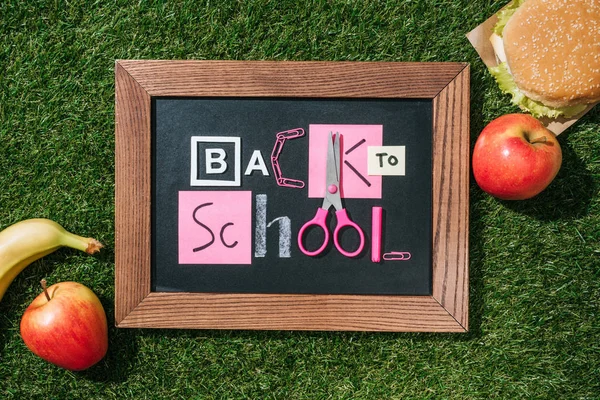 Tendido plano con manzanas frescas, plátano, hamburguesa y pizarra con letras de vuelta a la escuela de color rosa y blanco objetos en el césped verde - foto de stock