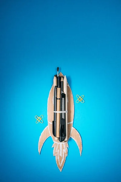 Top view of handmade cardboard rocket with stationery and paper clips on blue background — Stock Photo