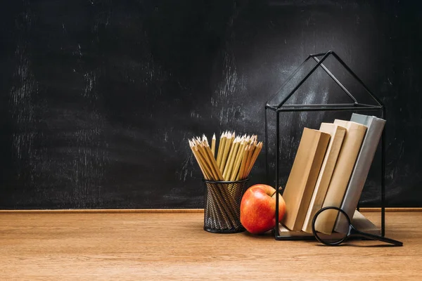 Close up view of fresh apple, magnifying glass, pencils and books on wooden surface with empty blackboard behind — Stock Photo
