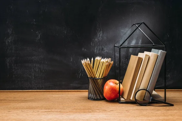 Vista de cerca de manzana fresca, lupa, lápices y libros en la superficie de madera con pizarra vacía detrás - foto de stock