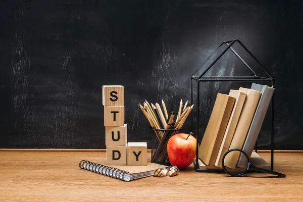 Close up view of study inscription made of wooden blocks, notebook, fresh apple and books on surface with empty blackboard behind — Stock Photo