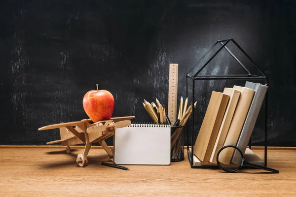 Close up view of apple on wooden toy plane, notebook, books and pencils on tabletop with empty blackboard behind — Stock Photo