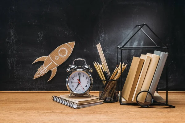 Primer plano vista de cohete de cartón en pizarra, lápices, reloj, cuaderno y libros en la mesa de madera - foto de stock