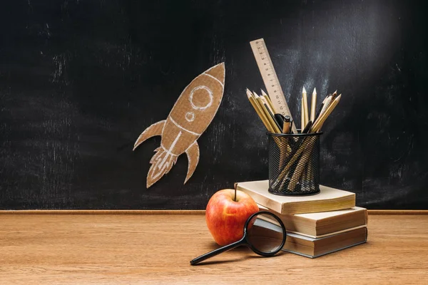 Vue rapprochée de fusée en carton sur tableau noir, pomme, loupe et livres sur plateau en bois — Photo de stock