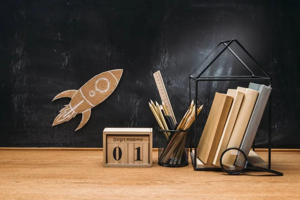 Close up view of cardboard rocket on blackboard, calendar, magnifying glass and books on wooden tabletop — Stock Photo