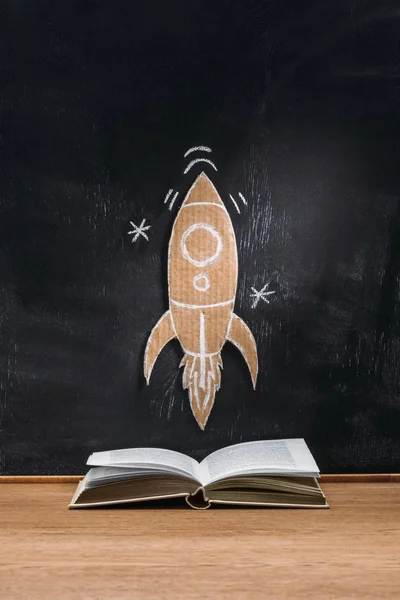 Vue rapprochée de fusée en carton sur tableau noir et livre ouvert sur surface en bois — Photo de stock