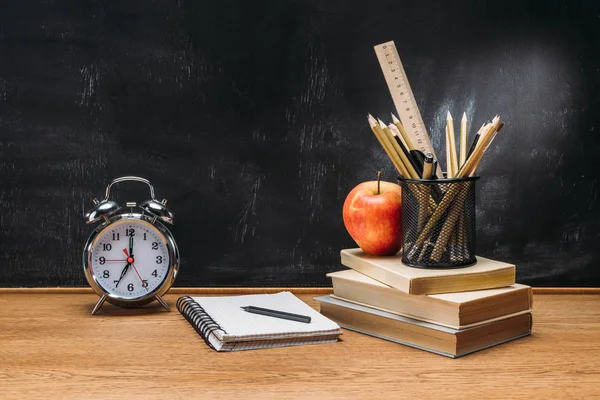 Close up view of fresh apple, clock, notebook, pencils and books on wooden tabletop with empty blackboard behind — Stock Photo