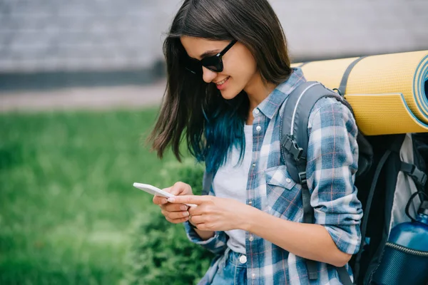 Улыбающаяся молодая женщина с рюкзаком с помощью смартфона — стоковое фото