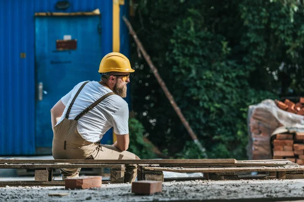 Rear view of young construction worker in hardhat and protective googles resting on pallet — Stock Photo