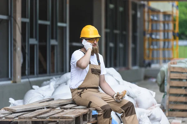 Jovem trabalhador da construção civil em chapéu duro e óculos de proteção falando no smartphone e almoçando com sanduíche — Fotografia de Stock