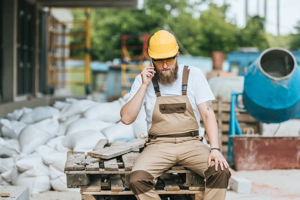 Builder in hardhat and protective googles smoking cigarette and talking on smartphone at construction site — Stock Photo