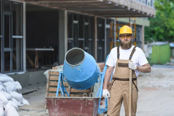 Jovem construtor em chapéu duro e óculos de proteção de pé com pá perto do misturador de concreto no local de construção — Fotografia de Stock