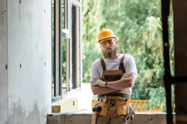 Lächelnder Bauarbeiter mit Schutzbrille und Hut, der mit verschränkten Armen auf der Baustelle steht — Stockfoto