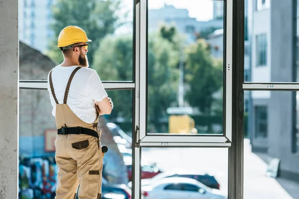 Rear view of builder in protective googles and hardhat standing with crossed arms and looking at windows — Stock Photo