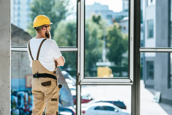 Visão traseira do construtor em óculos de proteção e hardhat de pé com braços cruzados e olhando para as janelas — Fotografia de Stock