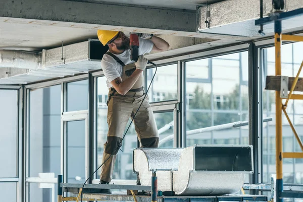 Constructor en googles de protección y perforación hardhat en el sitio de construcción - foto de stock