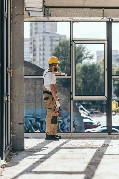 Vista trasera del constructor en hardhat y googles protectores instalando ventanas en el sitio de construcción - foto de stock