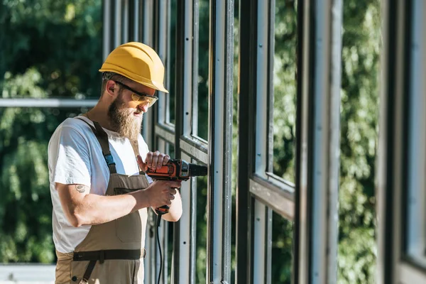 Vue latérale du constructeur en casque rigide et lunettes de protection installant des fenêtres par forage sur le site de construction — Photo de stock