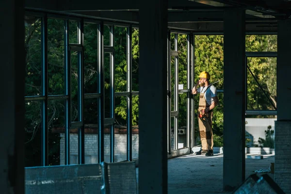 Vue à distance du constructeur en casque de protection et googles parler sur smartphone sur le site de la concstruction — Photo de stock