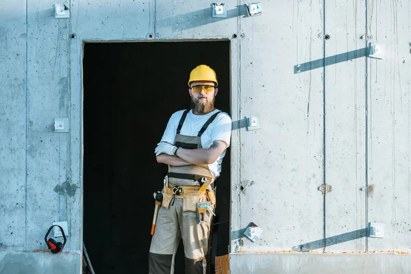 Builder in protective googles and hardhat standing with crossed arms at construction site — Stock Photo