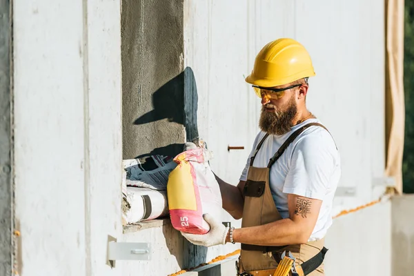 Joven constructor en googles protectores y bolsa de transporte de hardhat de cemento en el sitio de construcción - foto de stock