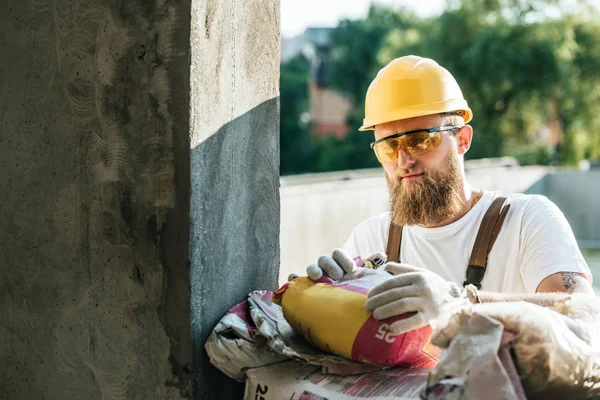Construtor em óculos de proteção e saco de abertura hardhat de cimento no local de construção — Fotografia de Stock