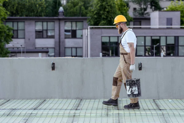 Seitenansicht des Bauarbeiters mit Schutzhelm und Schutzbrille, der auf der Baustelle Eimer mit Zement trägt — Stockfoto